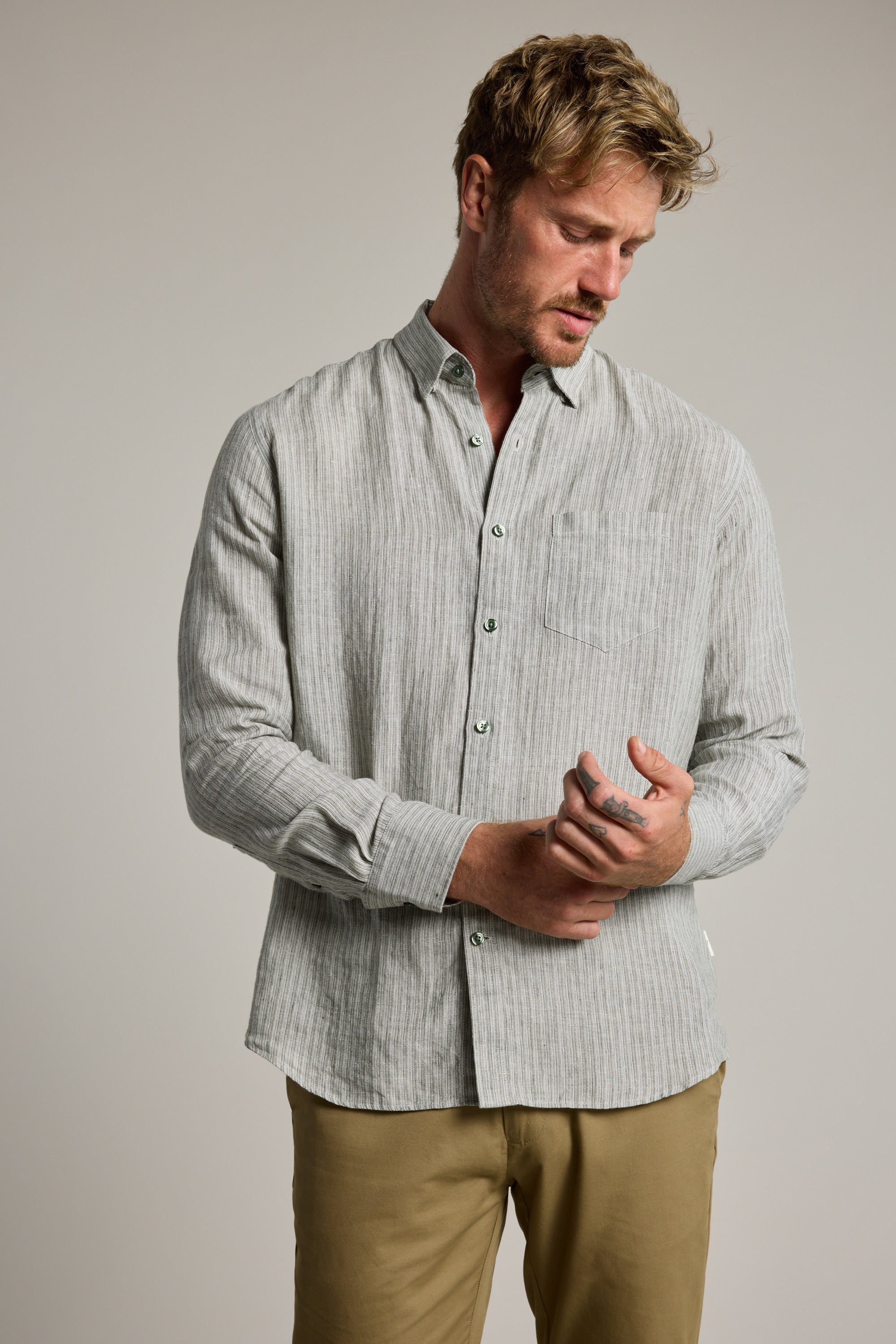 A man with light brown hair and a short beard wears the Barkers Dalton fine stripe Shirt in light gray and tan pants. He looks down, adjusting his left cuff with his right hand against a plain background.