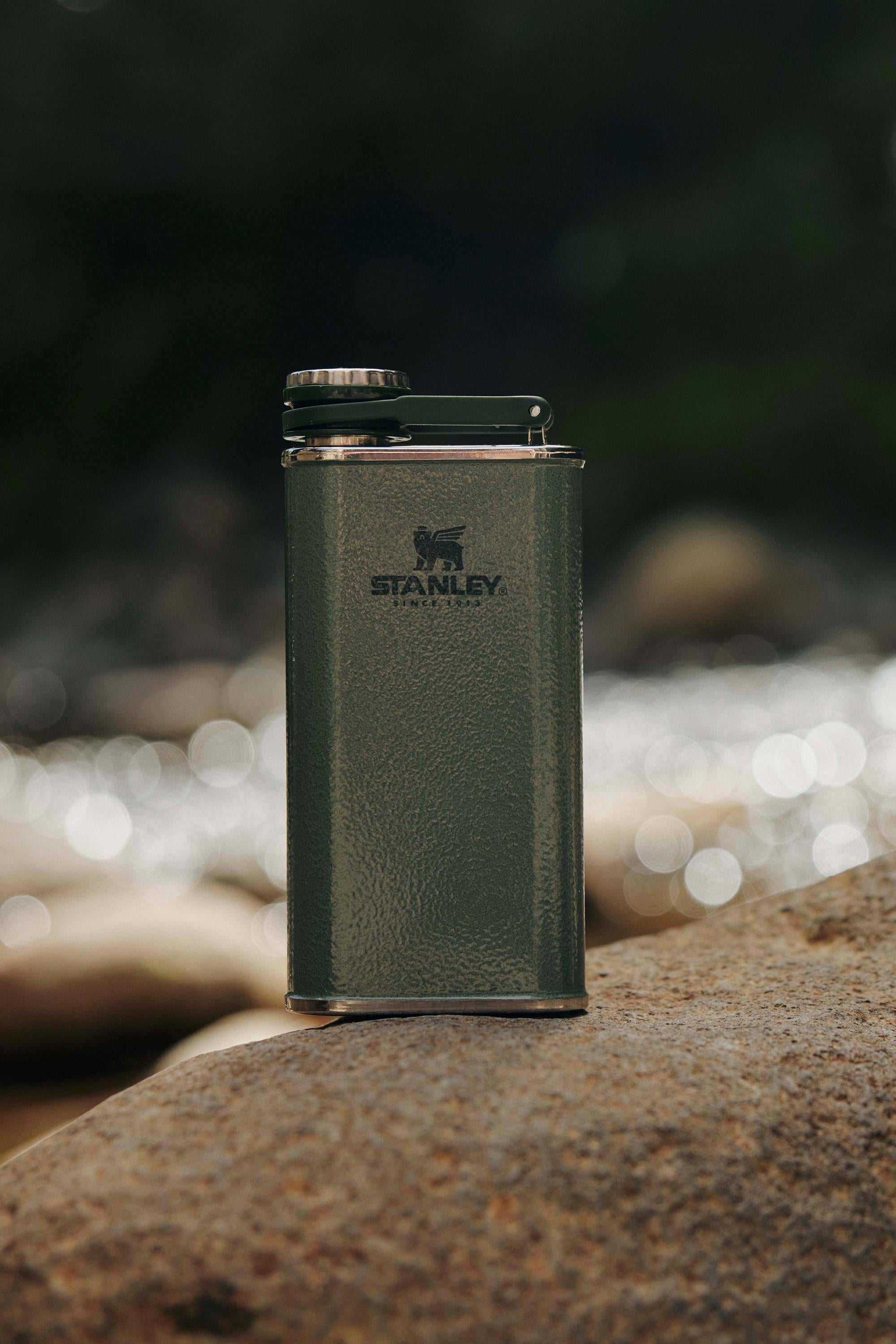 The Stanley Classic Easy Fill Wide Mouth Flask (235ml) by Stanley rests upright on a rock outdoors, making it an ideal adventure companion against a scenic backdrop of water and rocks.