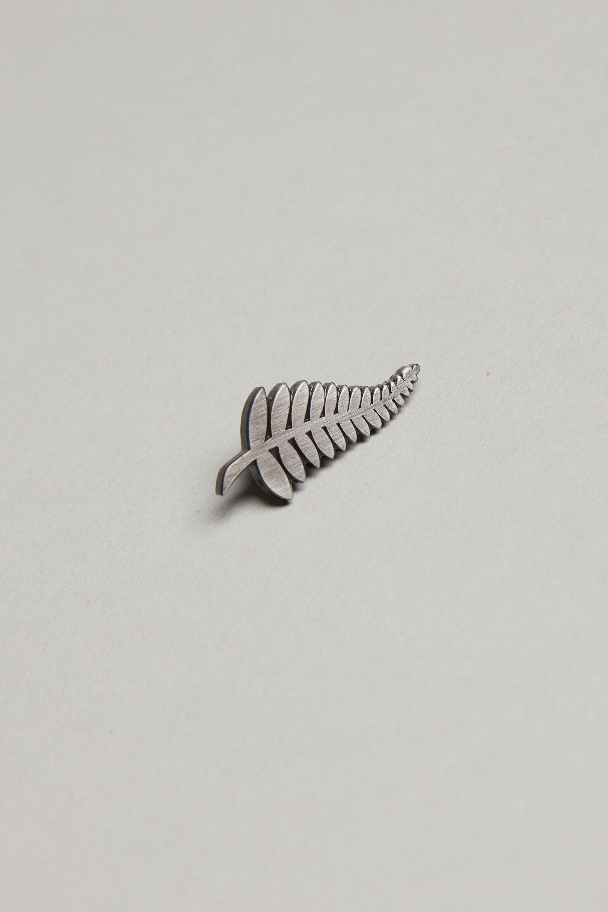 The Barkers Classic Fern Lapel Pin, featuring a brushed silver finish, rests on a plain light gray background—an ideal accessory to elevate any formal outfit.