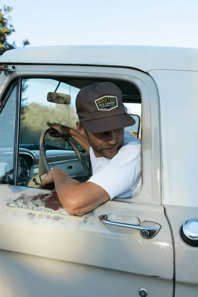 A man in a Brixton Coleson HP Snapback and white t-shirt sits in a vintage pickup truck, glancing over his shoulder with one hand on the wheel. The truck door is rusty, displaying the words "Custom Cab.