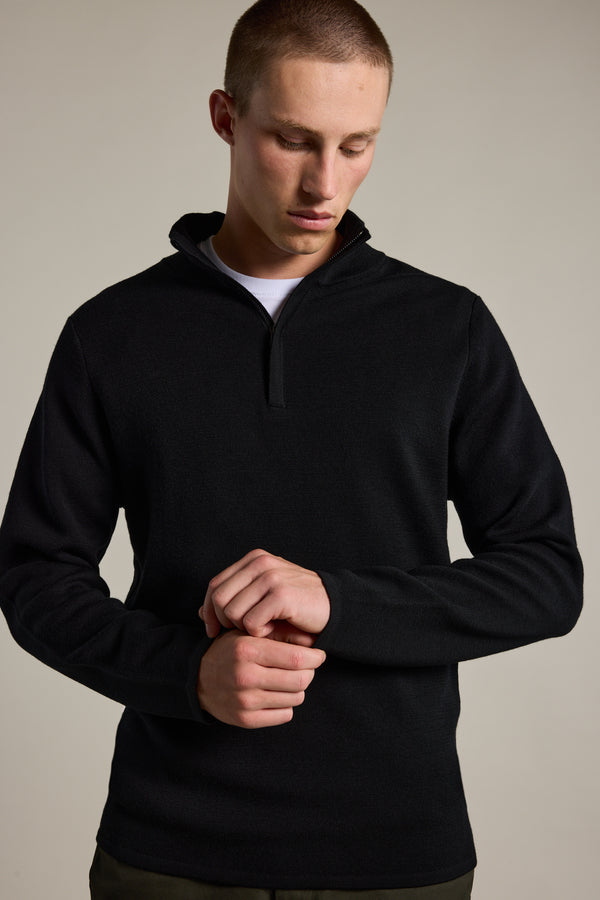 A person wears the Barkers Brandon ZQRX Merino 1/4 Zip pullover in black over a white shirt, standing against a plain background and adjusting their sleeve—ideal for cold-weather layering.