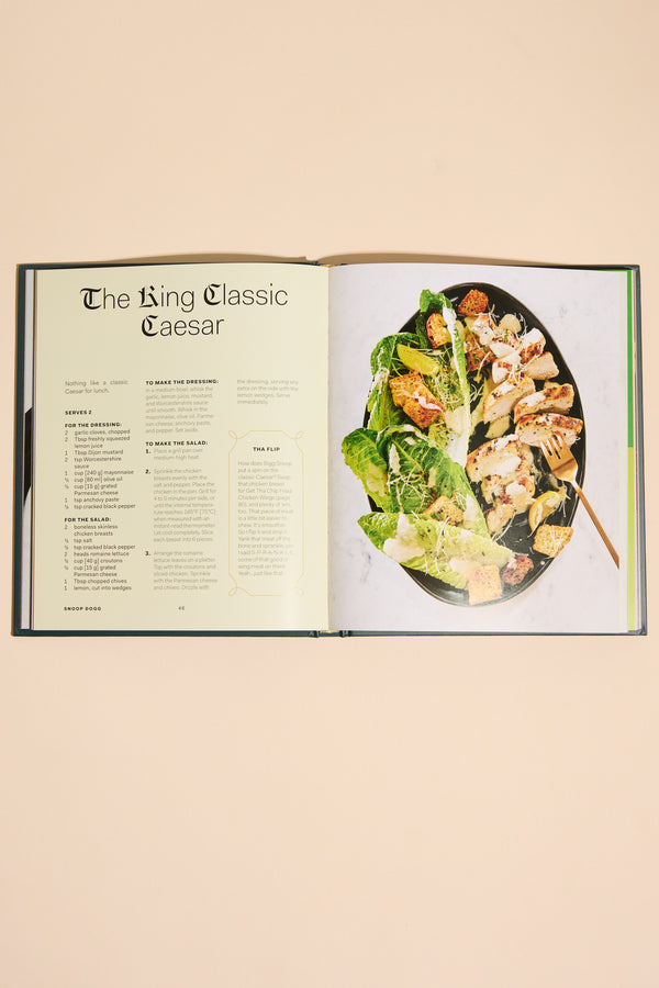 An open copy of "From Crook to Cook" by Other shows a cannabis-free King Classic Caesar recipe on the left and a photo of Caesar salad with lettuce, croutons, and dressing on a black plate with a fork on the right.