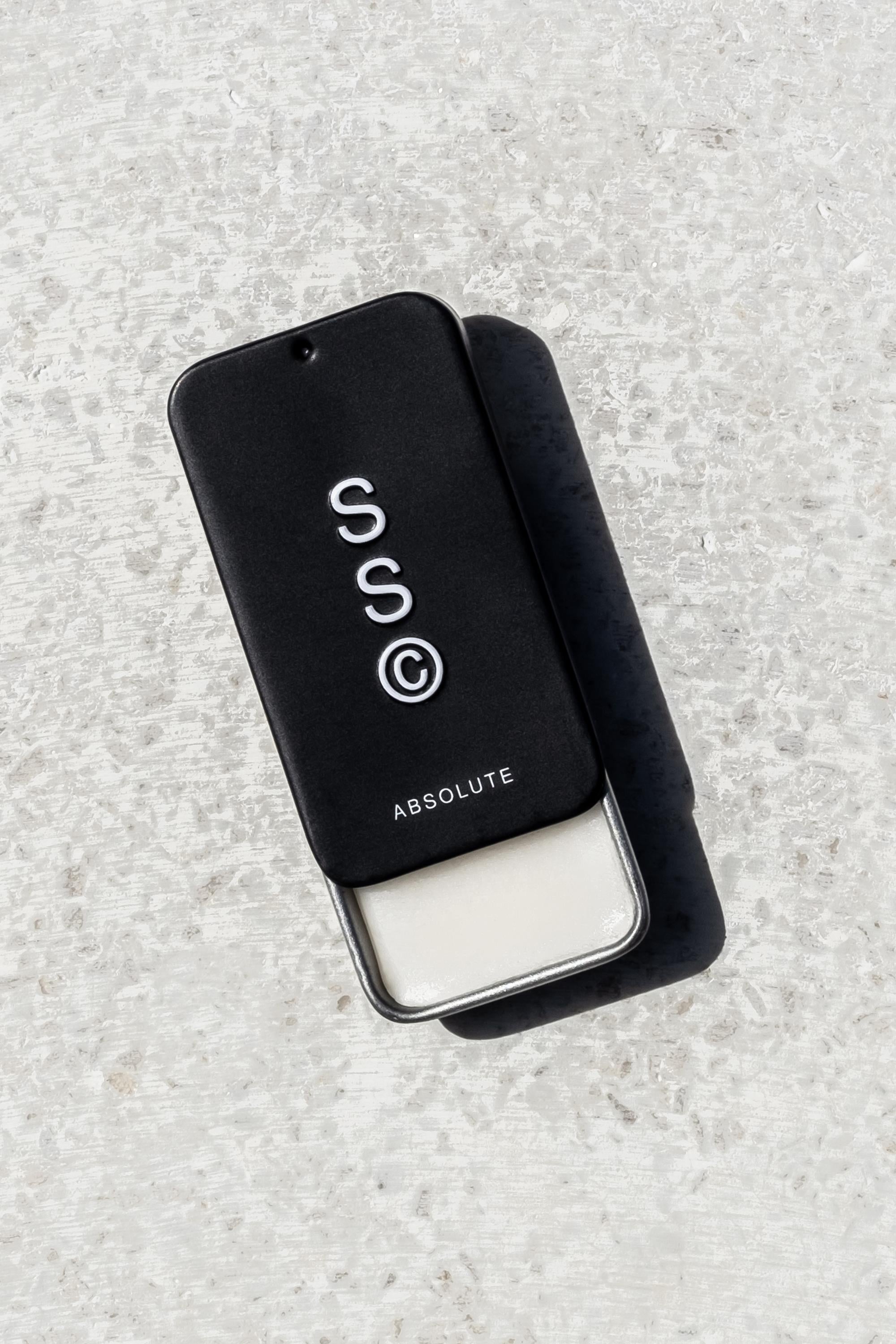 A black rectangular sliding tin labeled "Solid State - Absolute" with white solid cologne sits on a light surface, casting a shadow; this wax-based fragrance by Solid State is inspired by Creed Aventus.