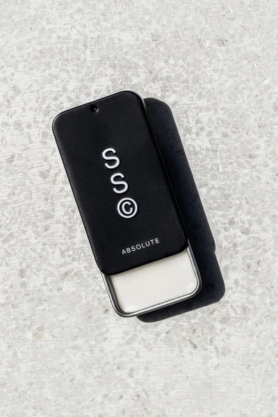 A black rectangular sliding tin labeled "Solid State - Absolute" with white solid cologne sits on a light surface, casting a shadow; this wax-based fragrance by Solid State is inspired by Creed Aventus.