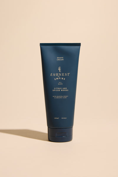 A dark blue tube of Other Earnest Empire Foaming Shave Cream in citrus and spiced woods scent stands upright on a beige surface with a beige background. Designed for sensitive skin, the label features gold and white text.