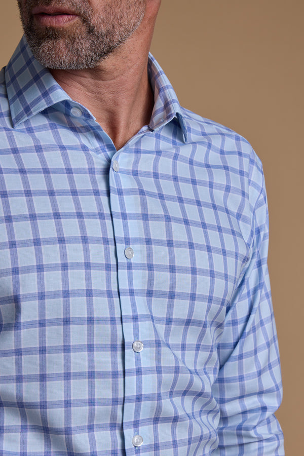 A man with a trimmed beard models the Barkers Bayard Check Business Shirt, a light blue and white plaid essential, against a beige background. Eco Easy Care fabric keeps him sharp as he turns his face slightly to the side.