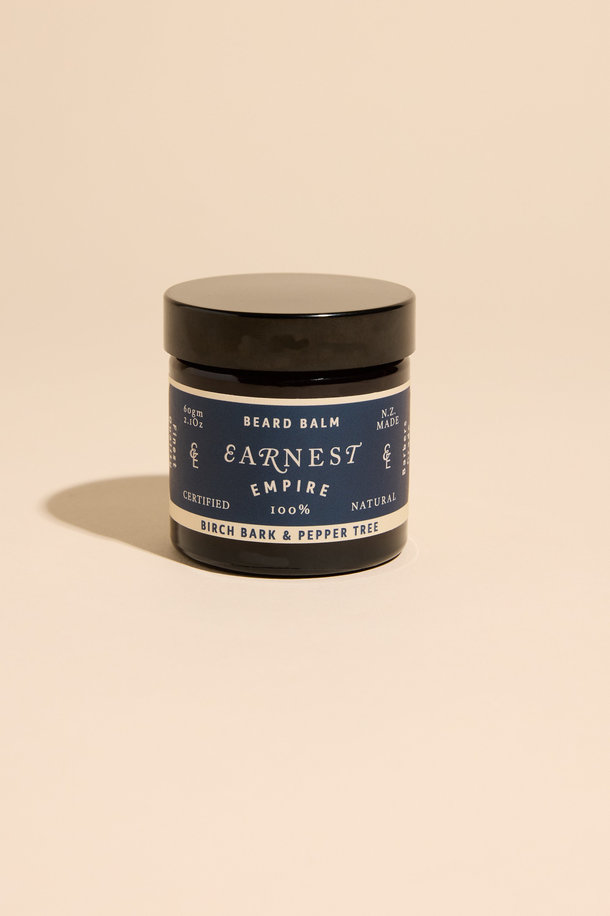 A black-lidded jar of Earnest Empire Beard Balm by Other rests on a beige backdrop. Its label features the Birch Bark & Pepper Tree scent and 100% natural ingredients, detailed in dark blue and white text.