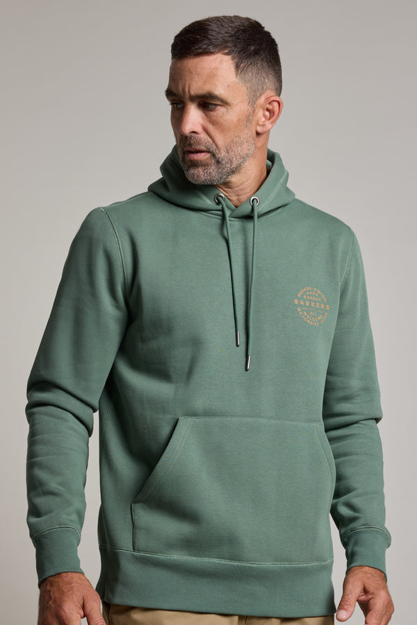 A man with short dark hair and a trimmed beard wears the Barkers Sunday Club Print Hoody—a bottle green cotton blend hoodie with a small circular logo on the left chest and front pocket—against a plain light grey background.