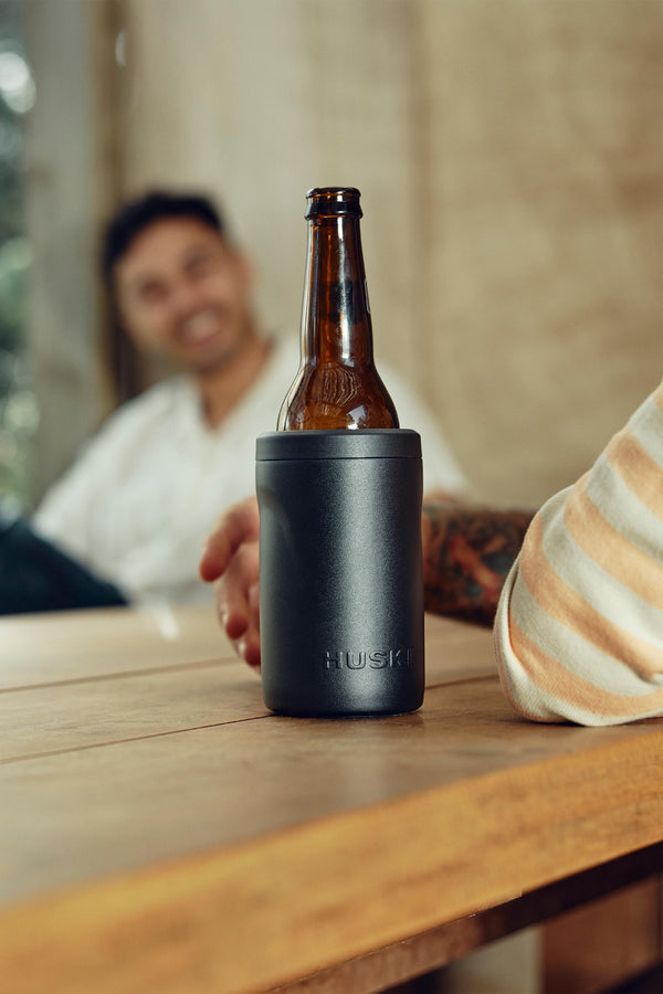 A close-up of a Huski Beer Cooler by Huski holding a brown beer bottle on a wooden table, with a smiling, blurred man in the background.