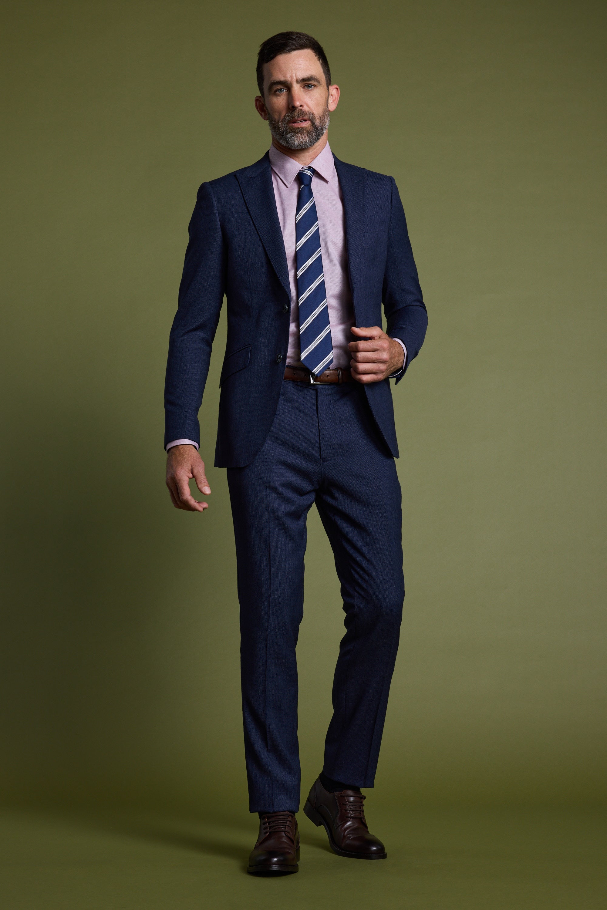 A man with short dark hair and a beard wears Barkers Watson Textured Suit Pants, a navy jacket, pink shirt, striped tie, and brown shoes, standing against an olive green background.