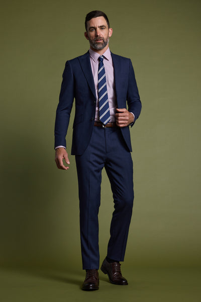 A man with short dark hair and a beard wears Barkers Watson Textured Suit Pants, a navy jacket, pink shirt, striped tie, and brown shoes, standing against an olive green background.