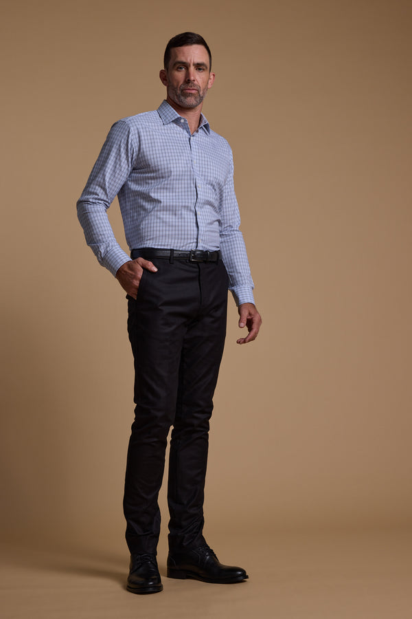 A man with short dark hair and a trimmed beard stands against a tan background, wearing a tailored fit Barkers Tabor Check Business Shirt in light blue, paired with black pants and dress shoes, one hand in his pocket.
