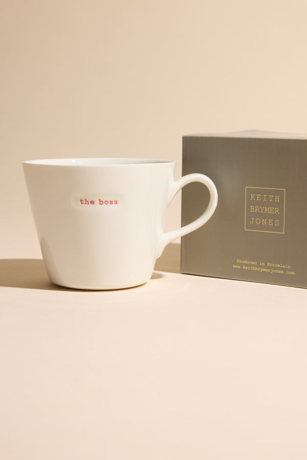 The Bucket Mug - The Boss by Other, featuring "the boss" in red on white ceramic, rests beside its gray box on a beige surface—a stylish homeware accent for neutral spaces.