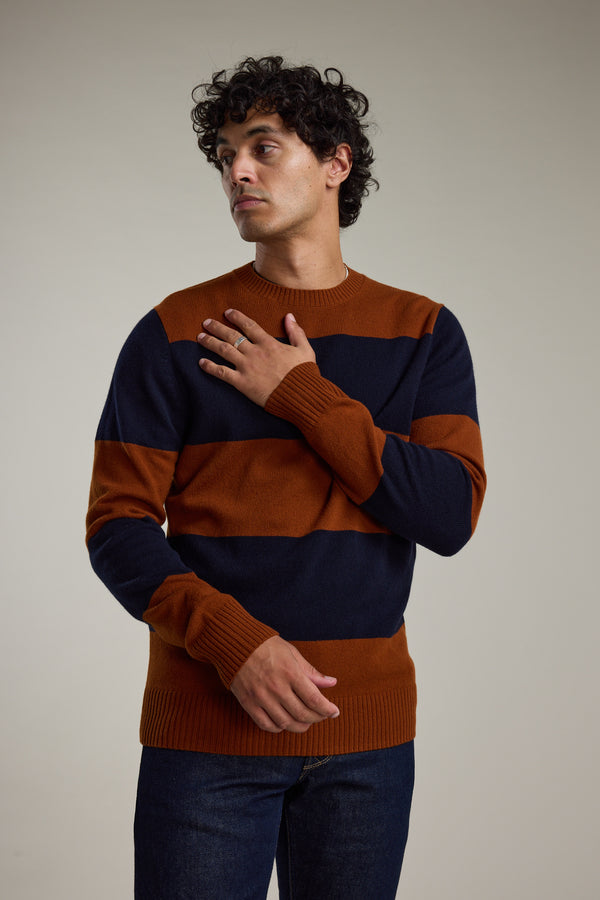 A man in the Barkers Portway Stripe Merino Crew stands against a plain background, gazing to the side with one hand on his chest and wearing dark jeans.