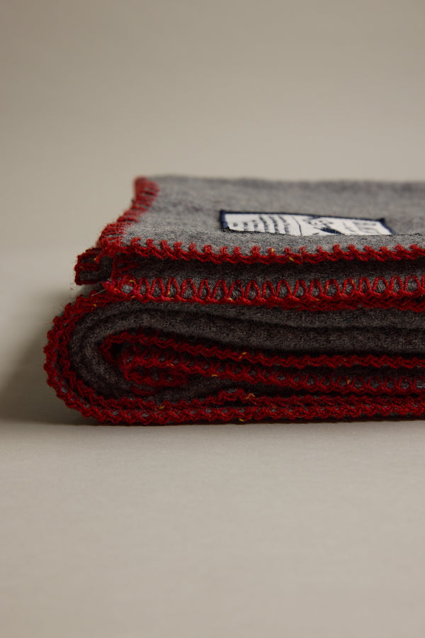 A folded gray Wool Take Back Blanket by Other, made from recycled wool with red edge stitching, lies on a light surface. The fabric label is visible on the top fold.