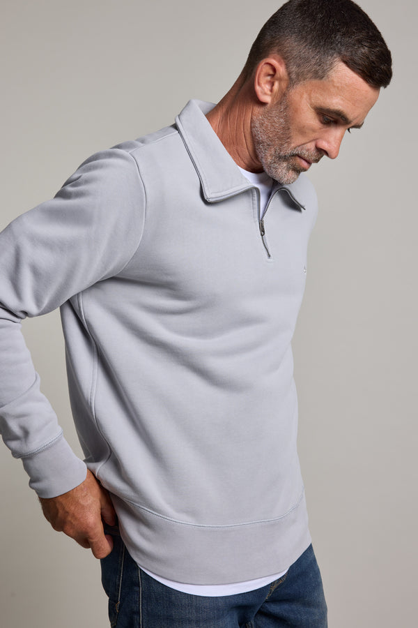 A man with short hair and a trimmed beard, wearing the Barkers Galway GD 1/4 Zip with a stand collar and blue jeans, stands against a plain light background, looking down with one hand in his pocket.