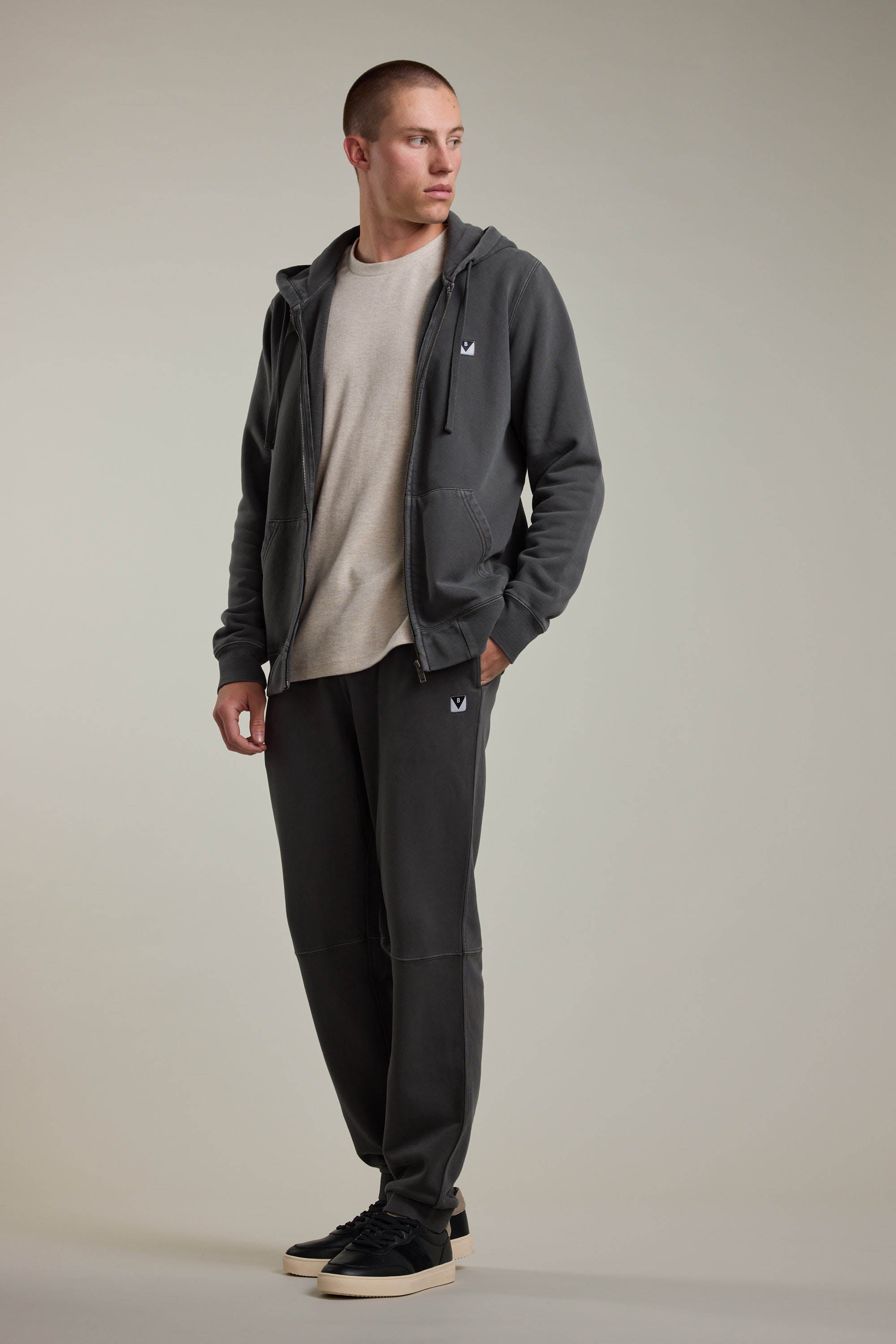 A person stands against a neutral background, wearing Barkers’ Newton GD Track Pant in a vintage wash, paired with a dark gray zip-up hoodie, light beige t-shirt, and black sneakers. One hand is in their pocket as they look slightly to the side.