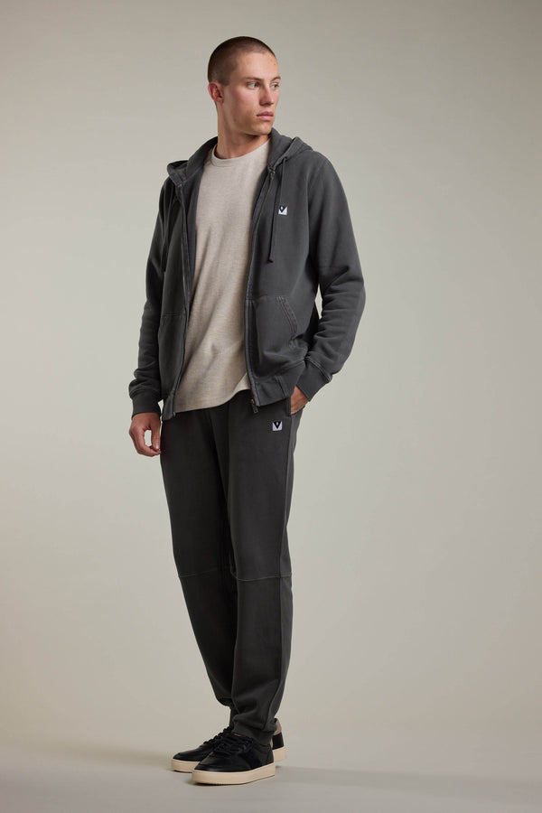 A person stands against a neutral background, wearing Barkers’ Newton GD Track Pant in a vintage wash, paired with a dark gray zip-up hoodie, light beige t-shirt, and black sneakers. One hand is in their pocket as they look slightly to the side.