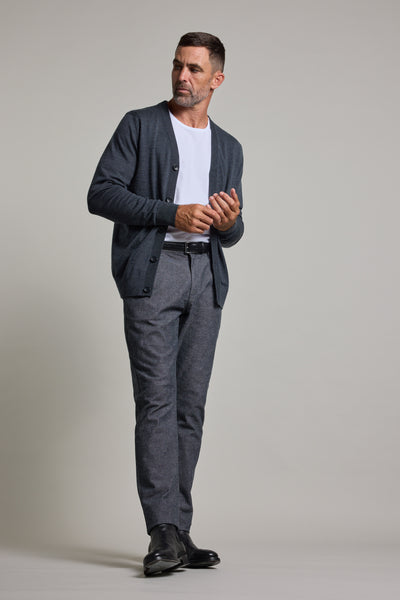 A man with short hair and a beard stands against a plain background, wearing the Barkers Skyland ZQrx Merino Cardi over a white t-shirt, paired with gray pants and black shoes. He looks to the side, hands loosely clasped.