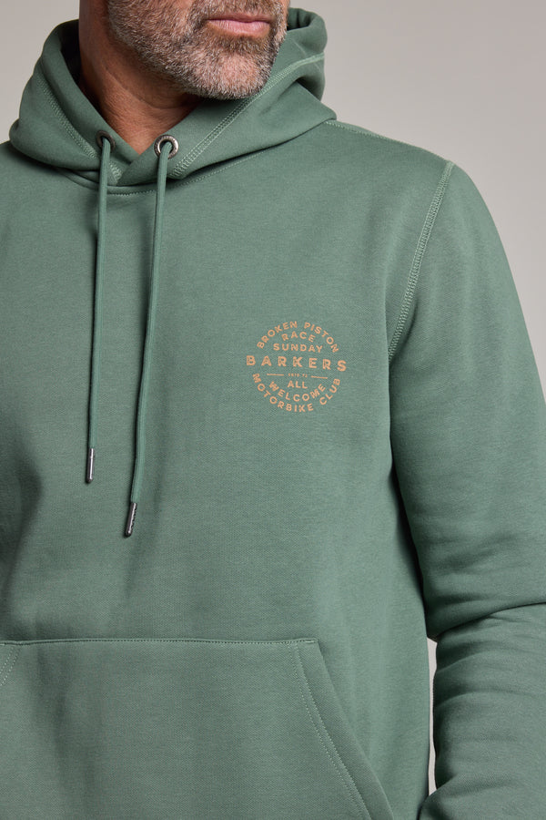 A man with a salt-and-pepper beard wears a sage green Barkers Sunday Club Print Hoody, featuring a circular orange "Boston, Sunday, Barbers, NYC, All Access, Brooklyn" logo on the left chest.