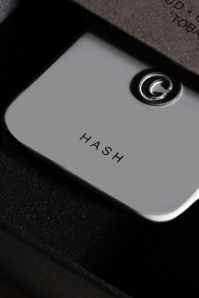 A close-up of Solid State - Hash, a sleek metallic solid state cologne by Solid State with a raised G logo, displayed in a black cushioned box.
