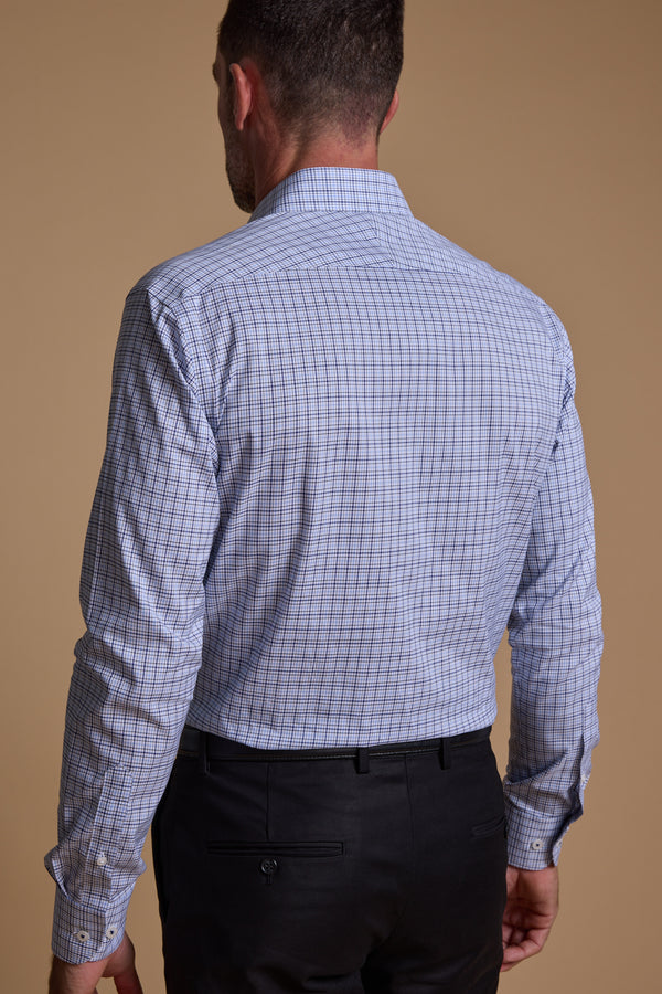 A man is seen from behind wearing the Barkers Tabor Check Business Shirt in light blue and white with a tailored fit and black trousers, standing against a beige background. This Eco Easy Care shirt blends comfort and style for any occasion.