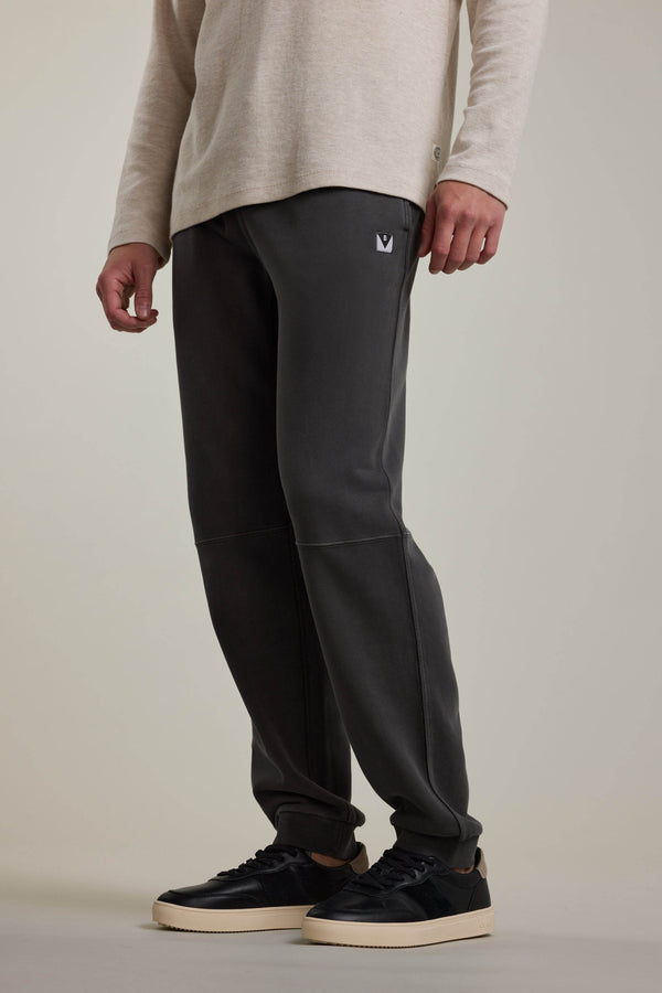A person wearing a light beige long-sleeve shirt, Barkers Newton GD Track Pant in dark gray with a small logo on the left thigh, and black sneakers with white soles, standing against a neutral background.