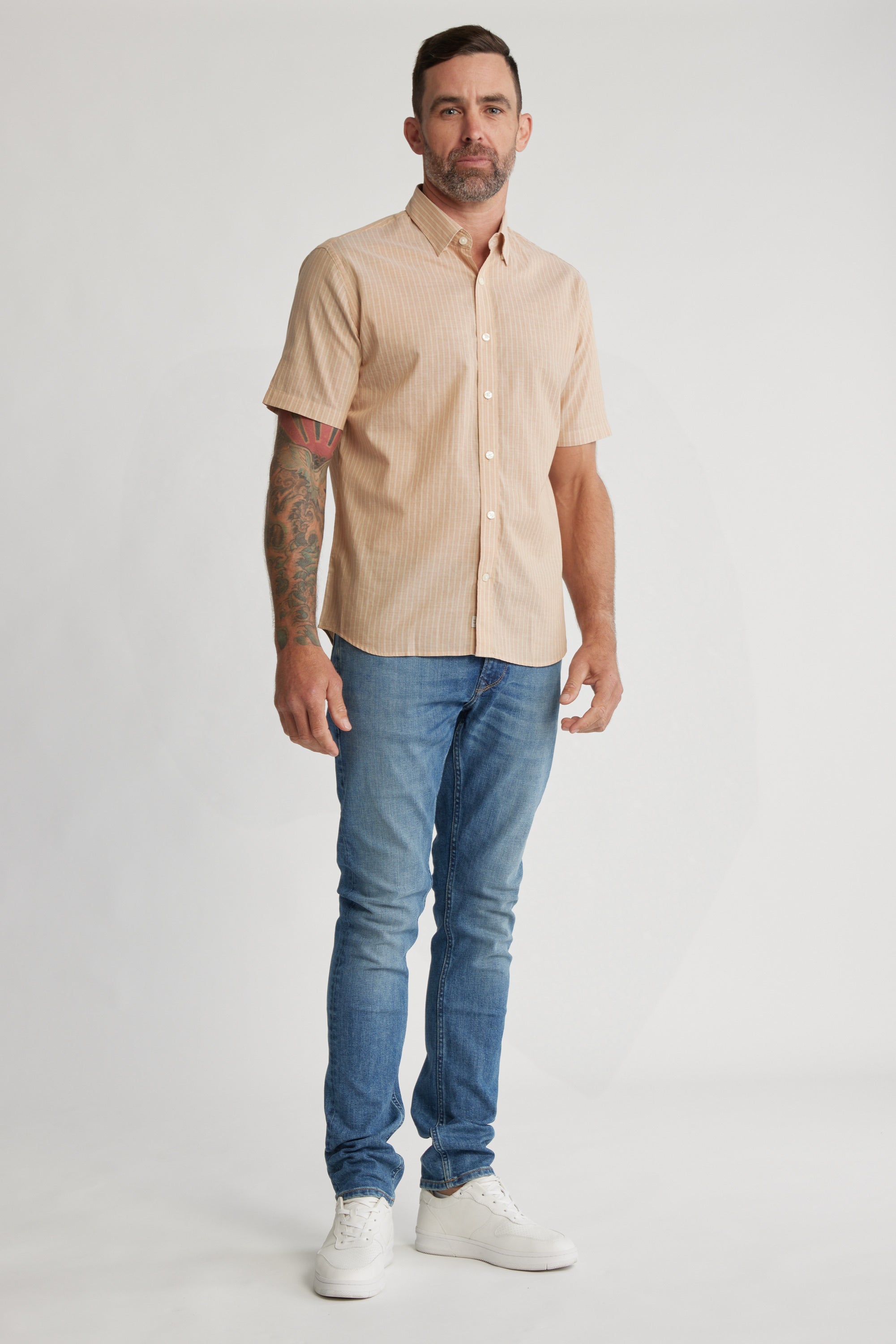 A man with short hair, a beard, and a tattooed arm stands against a plain background wearing the Barkers Burnham Stripe Shirt, blue jeans, and white sneakers.