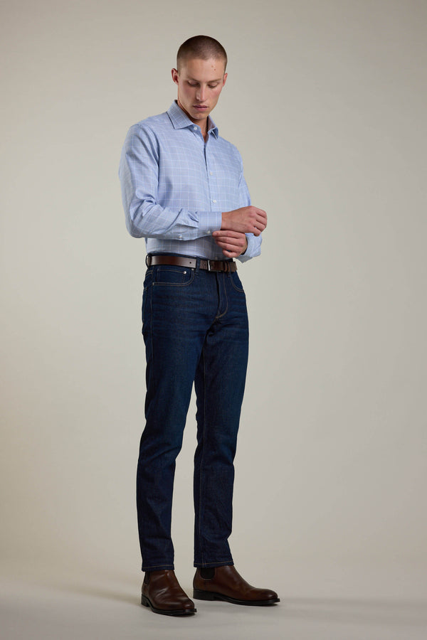 A man stands against a plain background wearing the Barkers Tyson Check Business Shirt. He pairs it with dark blue jeans, a brown belt, and brown shoes while looking down and adjusting his shirt cuff with one hand.