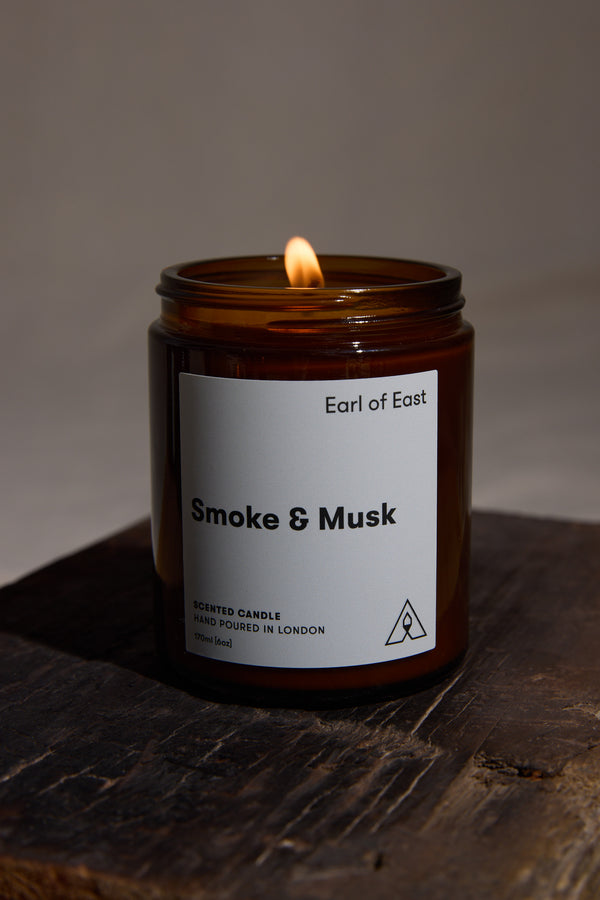 The Earl of East EoE Smoke And Musk Candle 170ml, crafted from soy wax, sits on a dark wooden surface with a soft neutral background. Earthy patchouli notes complement its warm glow through brown glass.