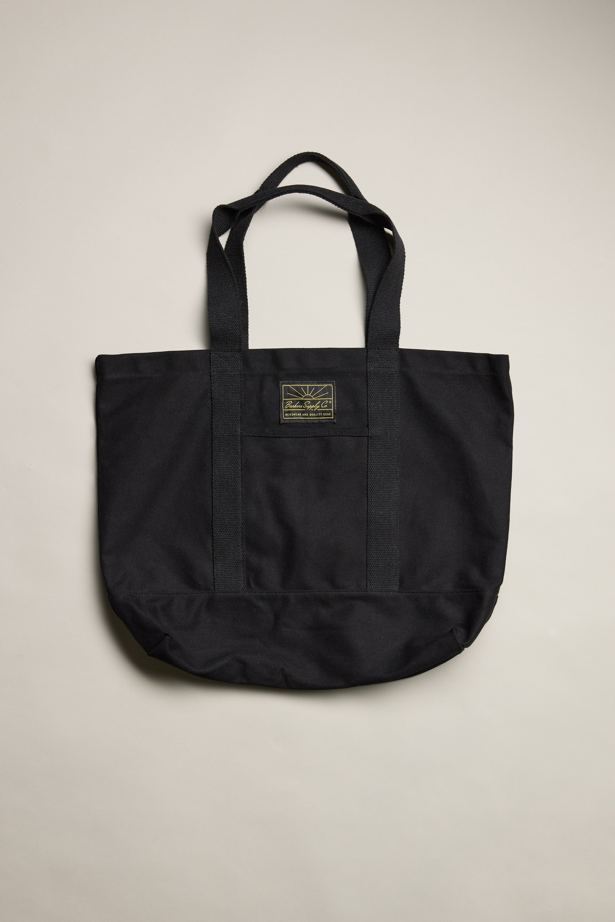 The Barkers Supply Co. Canvas Tote Bag by Barkers features two handles and a small rectangular patch label at the top center. Made from durable black canvas, it's displayed against a plain light background.