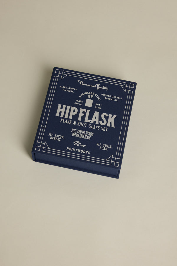 A navy blue gift box labeled “Barkers The Essentials Hip Flask” with white vintage-style text and decorative borders sits on a light beige surface, highlighting premium quality and durable stainless steel construction.