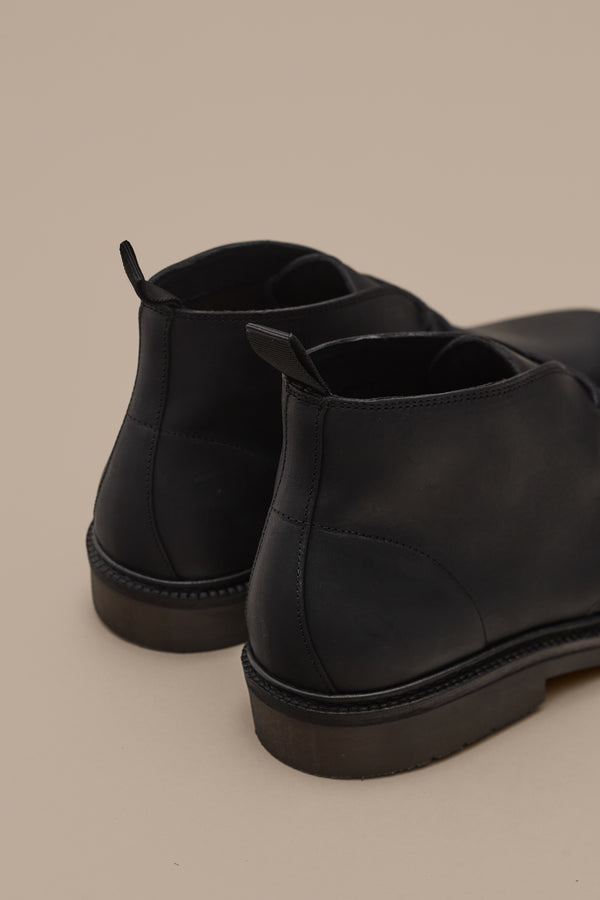 A close-up of the back and sides of Barkers' Howarth Desert Boot in black leather, featuring pull tabs and classic Portuguese-inspired design, displayed against a neutral beige background.