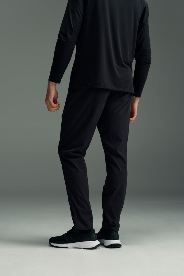 Photographed from behind on a light gray floor with a plain gray background, a person wears the WearEver Transit Pant by WearEver, paired with a long-sleeved black top and black sneakers.