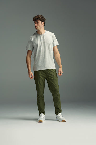 A man with curly hair stands against a plain grey background, wearing a light grey t-shirt, white sneakers, and WearEver’s tapered fit olive green Transit Pant, looking off to the side with a neutral expression.