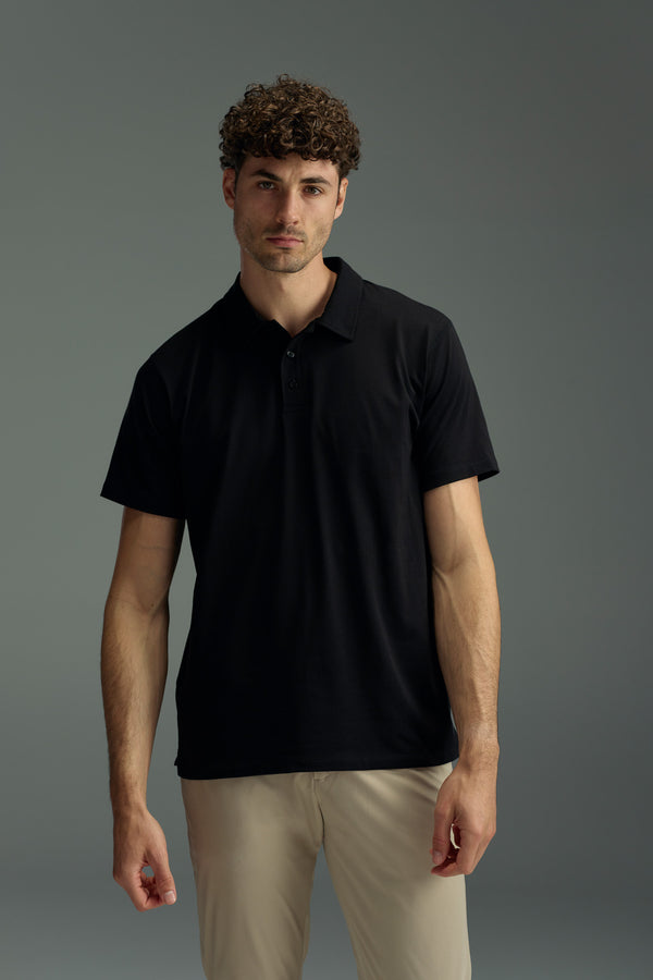 A man with short curly hair wears the black WearEver Outbound Polo from WearEver and beige pants, standing against a plain grey background and looking directly at the camera with a neutral expression.