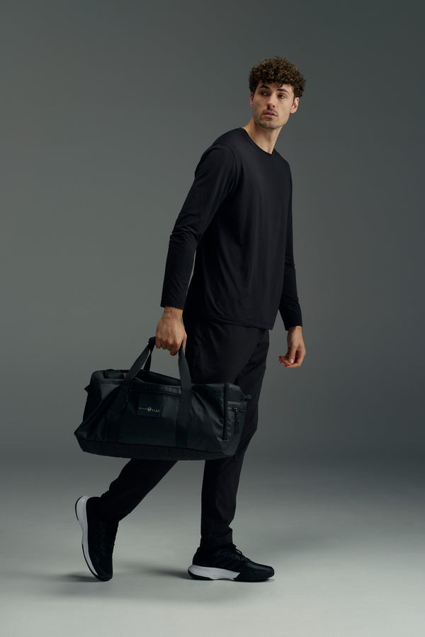 A man with curly brown hair, wearing a black long-sleeve shirt, black pants, and black sneakers, holds the WearEver Transit Holdall by WearEver in his left hand while looking right against a plain gray background.