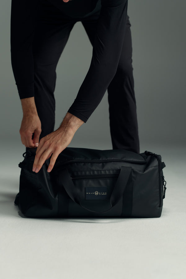 Dressed in black, a person bends over and zips the large, water-resistant WearEver Transit Holdall duffel bag, featuring a rectangular WearEver logo on the front, set against a plain light background.
