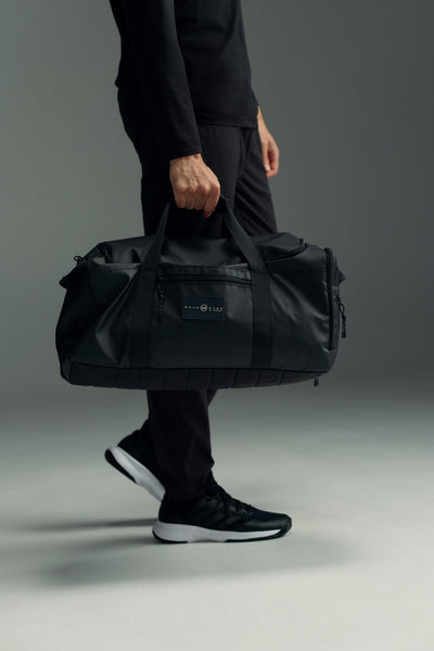 A person in black athletic wear holds the WearEver Transit Holdall, a black water-resistant duffel bag by WearEver, by its handles. Only their lower half is visible against a plain, neutral background.