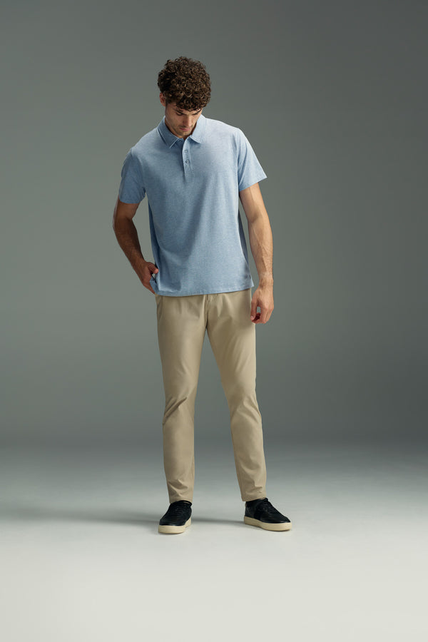 A man in a light blue WearEver Outbound Polo by WearEver, crafted from breathable cotton-blend fabric, pairs it with beige pants and black sneakers as he stands against a gray background, looking down with one hand in his pocket.