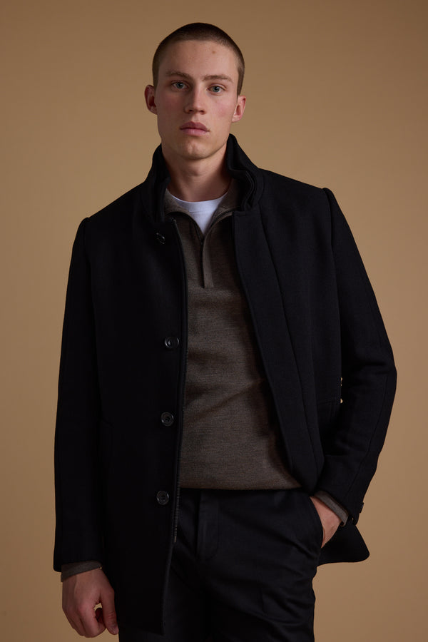 A young man with short hair stands against a beige background, wearing the Barkers Raymond Coat over a brown sweater and white shirt, one hand in his pocket, gazing at the camera with a neutral expression.