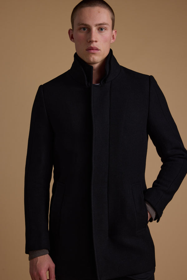 A person with short hair models the Barkers Raymond Coat, a high-collared black wool blend, standing against a plain beige background and looking directly at the camera with a neutral expression.