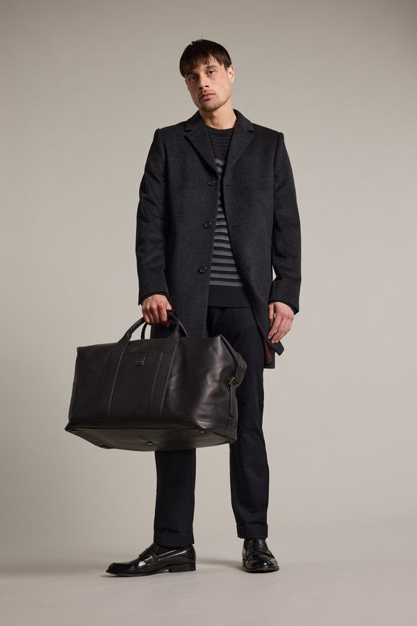 A man in a dark coat, striped sweater, and black pants stands holding the Barkers Costello Leather Weekender duffel bag at his side. With short brown hair and a neutral expression, he poses against a plain backdrop.
