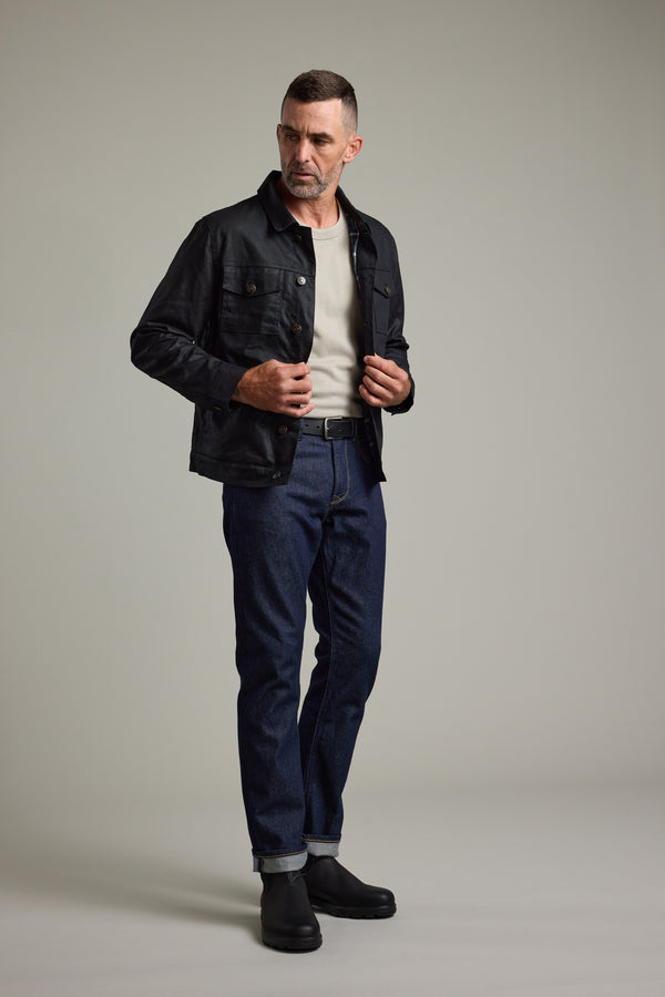 Man in a Barkers Benson Waxed Cotton Jacket, beige shirt, blue jeans, and black boots in studio.