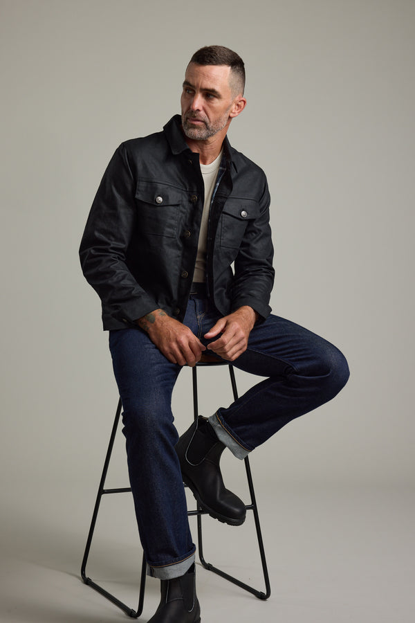 Man in Barkers Benson Waxed Cotton Jacket and jeans sits on a stool against a plain background.