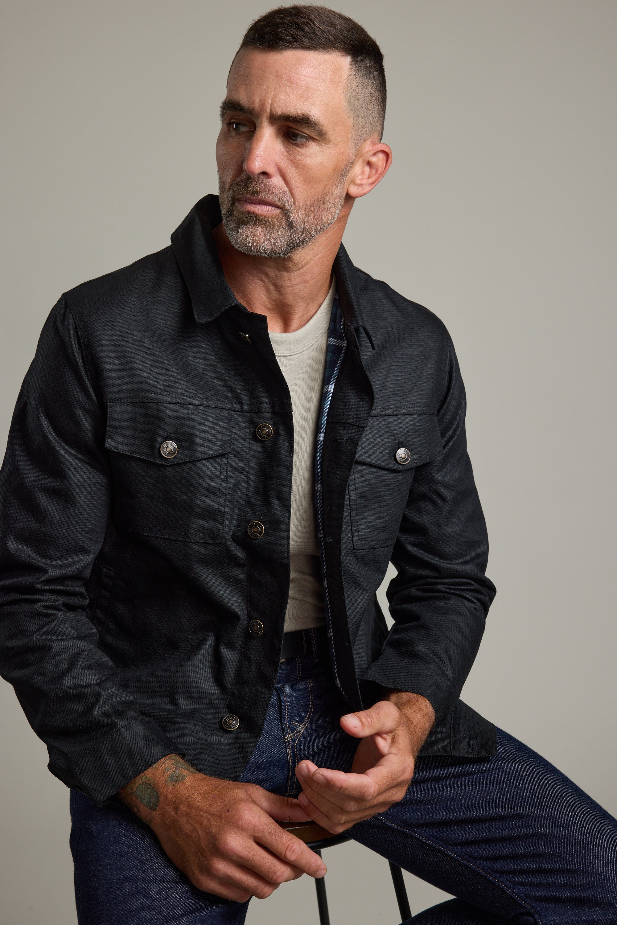 Man in Barkers Benson Waxed Cotton Jacket and jeans, sitting on a stool.
