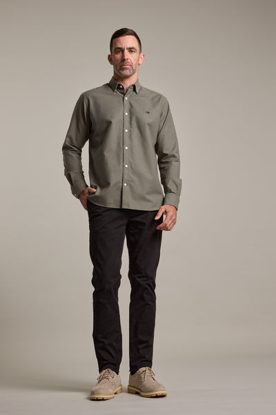 A man faces forward against a plain light background, wearing a button-up olive green shirt, Barkers Carnaby Chino in black, and beige shoes, with one hand in his pocket.