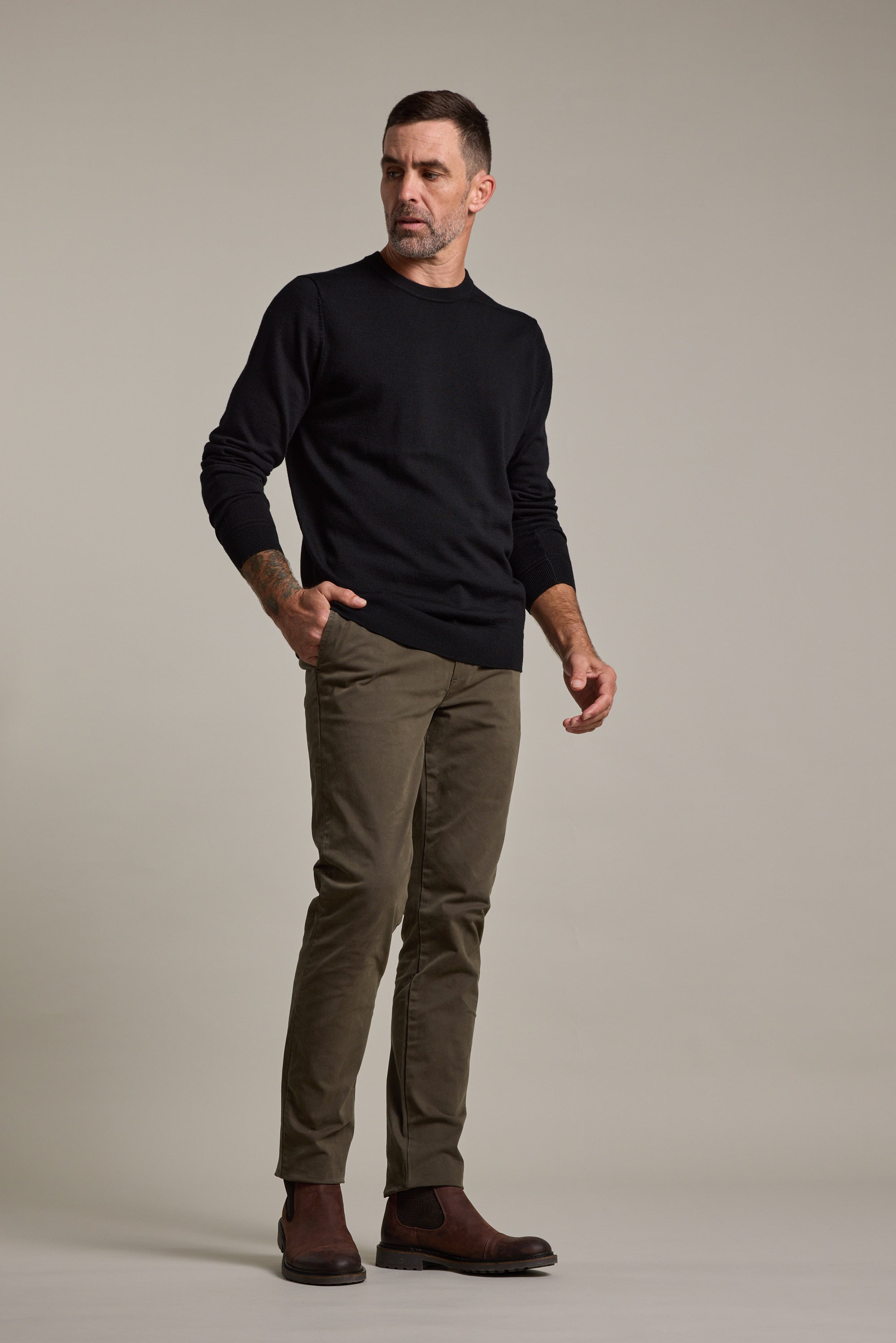 A man with short dark hair and a beard stands against a plain background, wearing a black long-sleeve sweater, Barkers Carnaby Chino in olive green, and brown boots. He looks slightly to the side with a neutral expression.