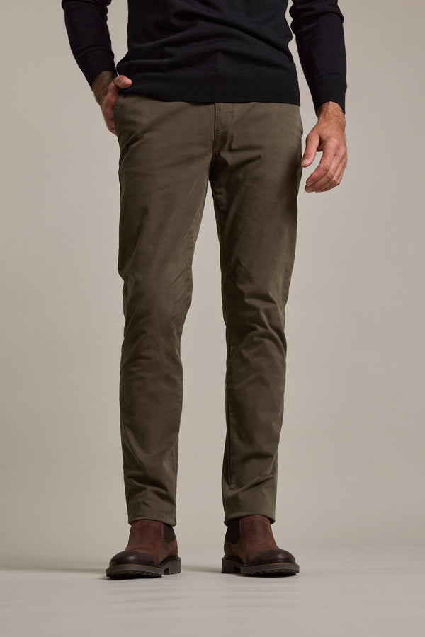 A person wears Barkers' Carnaby Chino in olive green, made from responsibly sourced cotton, styled with a black sweater and brown leather shoes. Only their lower body is visible against a plain light background.