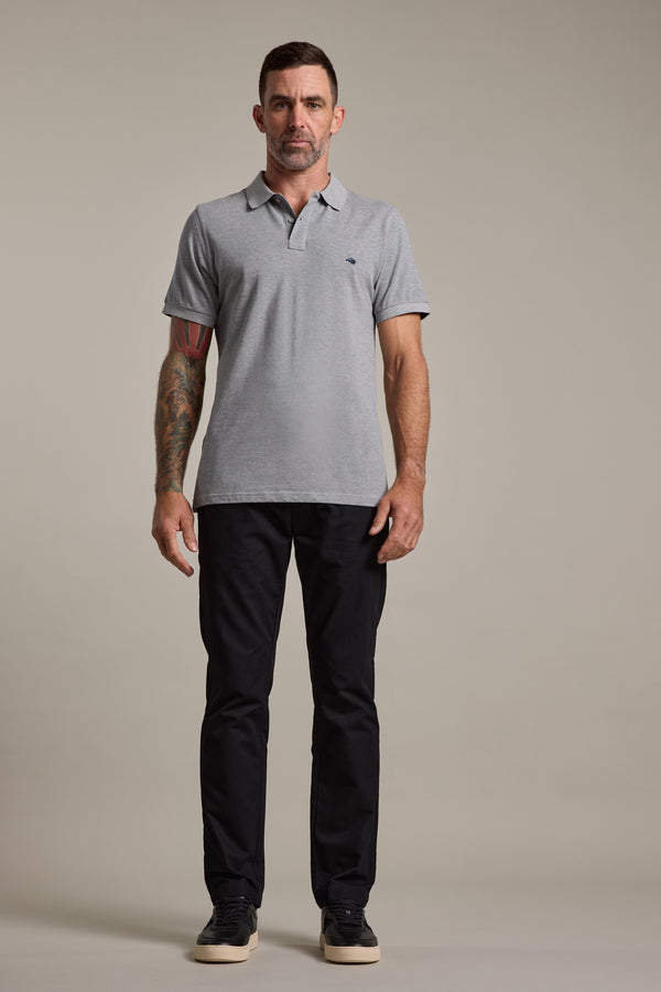 A man with short dark hair and a tattooed left arm stands facing forward, wearing a light gray polo shirt, Barkers Dundas Chino trousers in relaxed fit cotton twill, and black sneakers against a plain beige background.